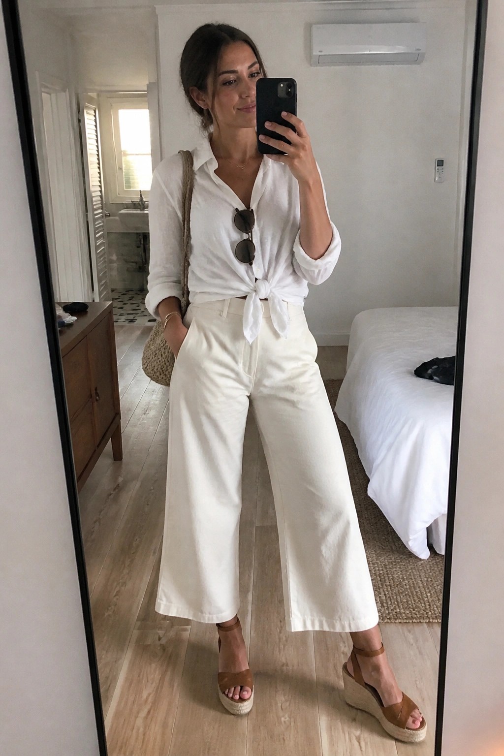 Woman in white knotted shirt and wide-leg pants taking bedroom mirror selfie with wedges