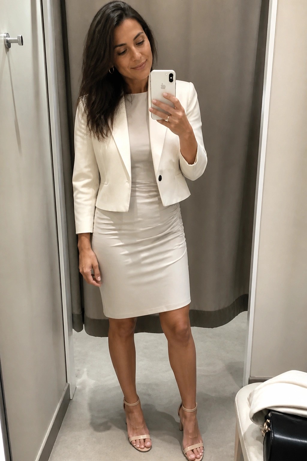 Woman in a white cropped blazer over a fitted white dress with beige ankle-strap heels holding a phone for a mirror selfie