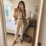 Woman in beige blazer and trousers taking mirror selfie in bedroom