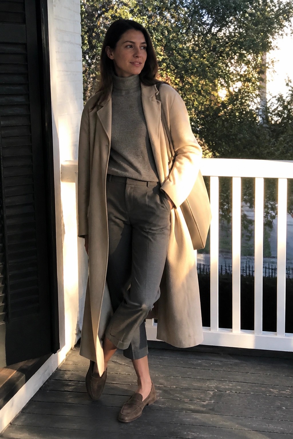 Woman in a long beige trench coat layered over a gray turtleneck sweater and matching cropped gray trousers paired with brown suede loafers.
