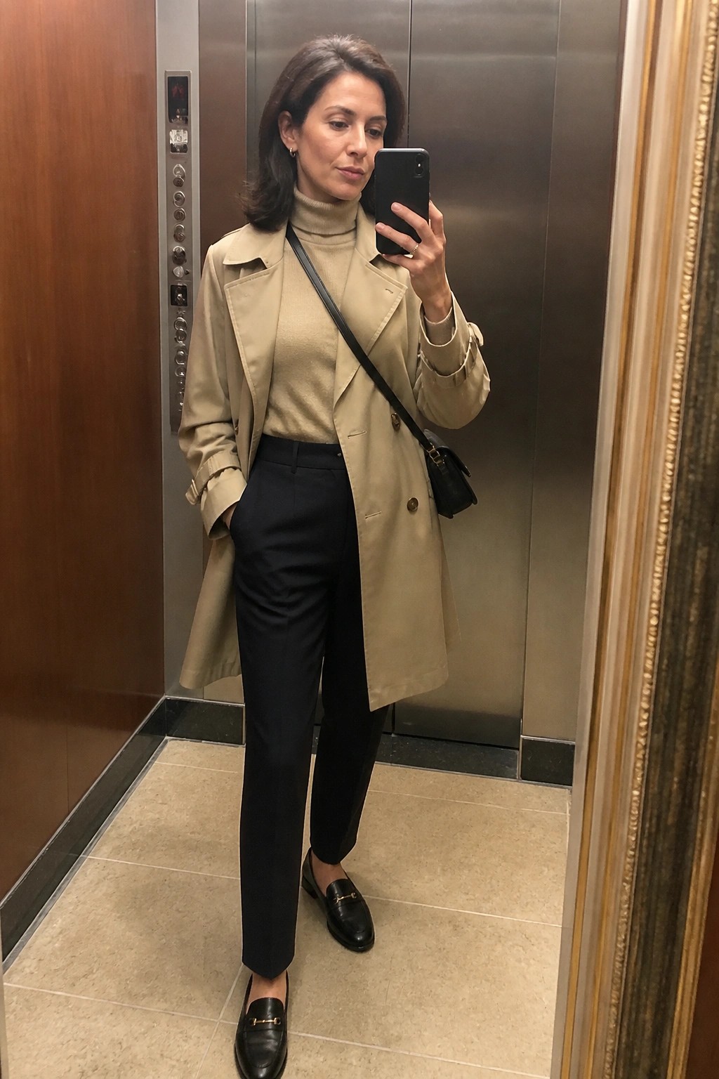 Woman in a beige trench coat over a beige turtleneck sweater paired with black tailored trousers and black loafers standing in an elevator taking a mirror selfie.