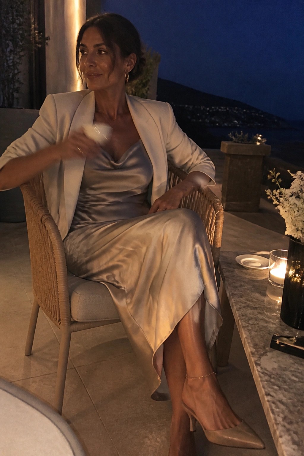 Woman seated in a light beige blazer over a silvery satin slip dress with cowl neck, paired with matching pointed toe heels and a delicate gold anklet.