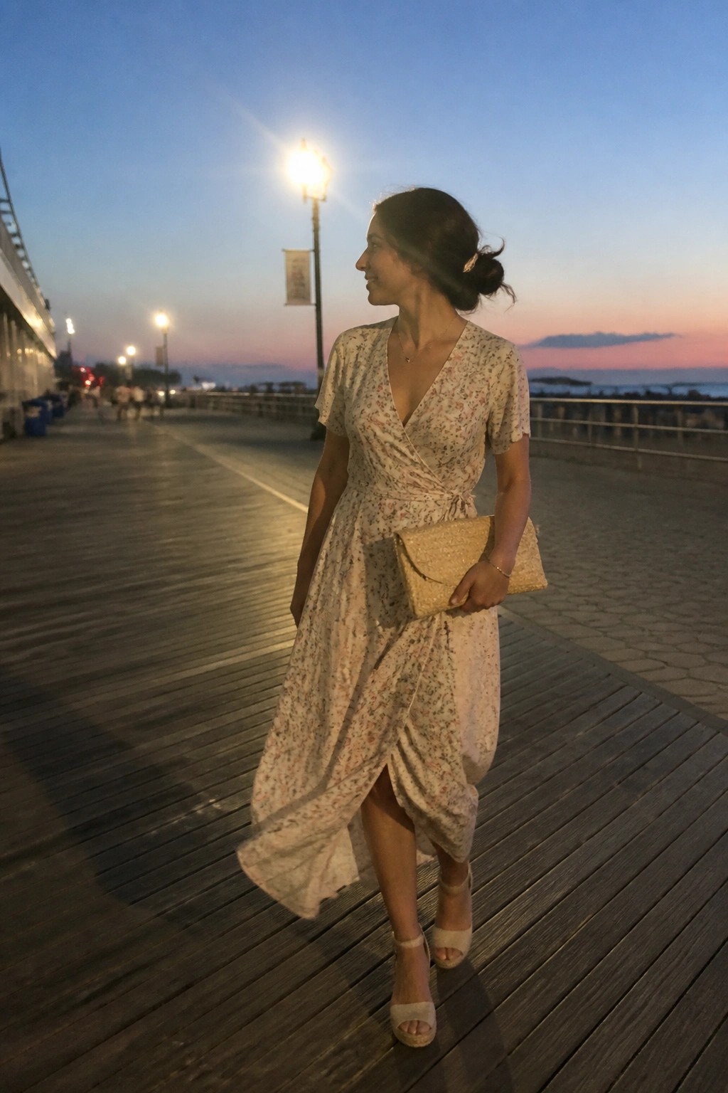 Woman wearing a cream floral print wrap dress with short sleeves, V-neckline, waist tie, and asymmetrical hem, paired with beige block heel sandals and a gold clutch.