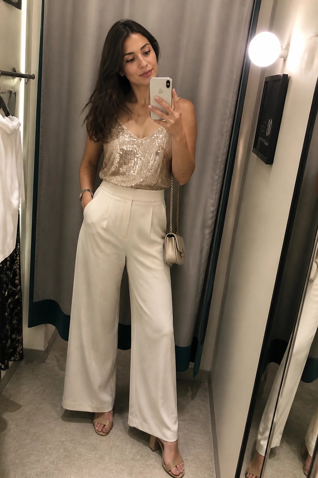 Woman in sequin top and white wide-leg pants taking mirror selfie in fitting room.