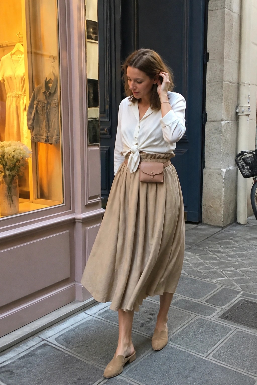 Woman in white tied shirt and beige midi skirt with pouch bag on cobblestone street