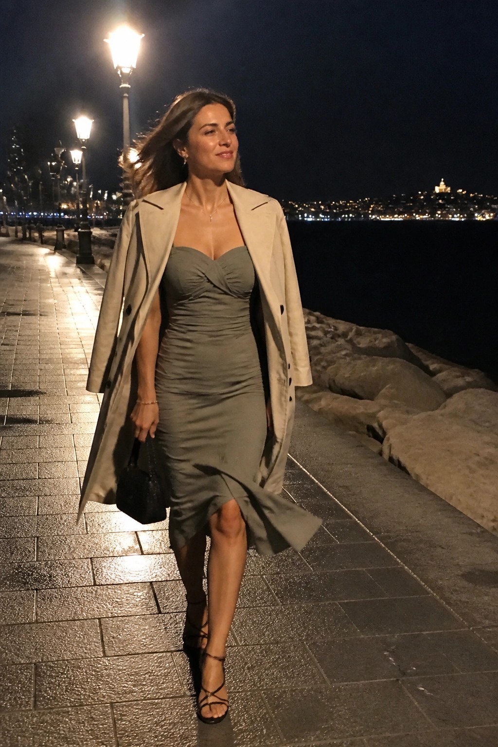 Woman wearing sage green ruched midi dress under open beige trench coat with black strappy heels and small black handbag.