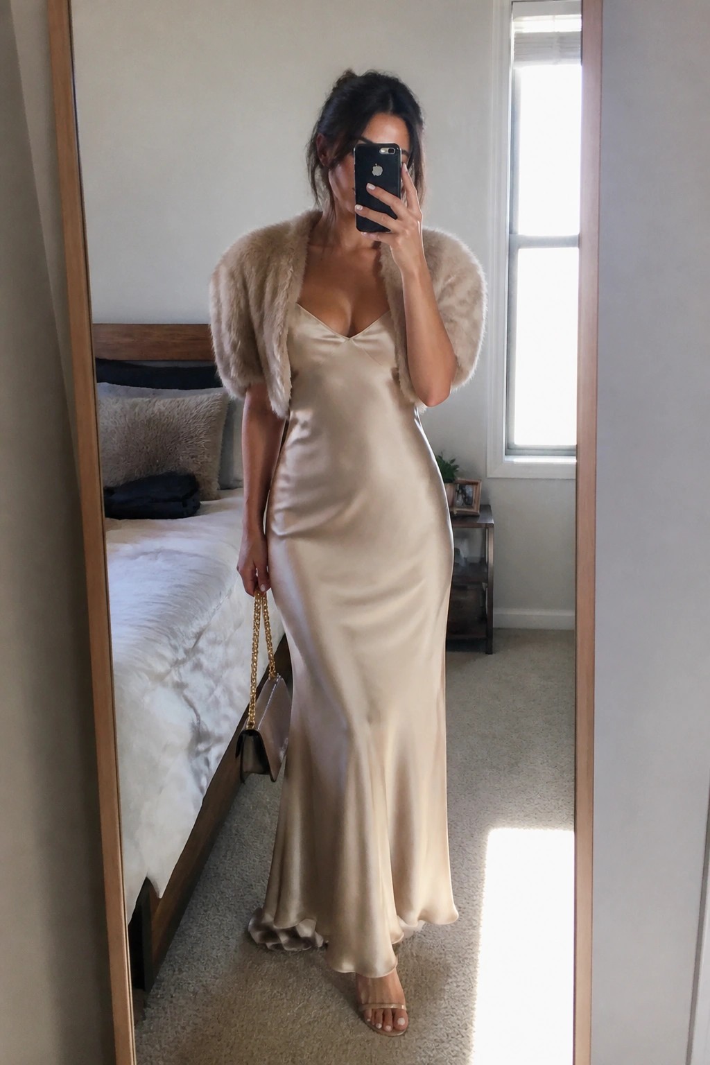 Woman wearing a floor length champagne satin slip dress with V neckline and a short beige fur bolero jacket, holding a small taupe handbag with gold chain strap, standing in front of a mirror.