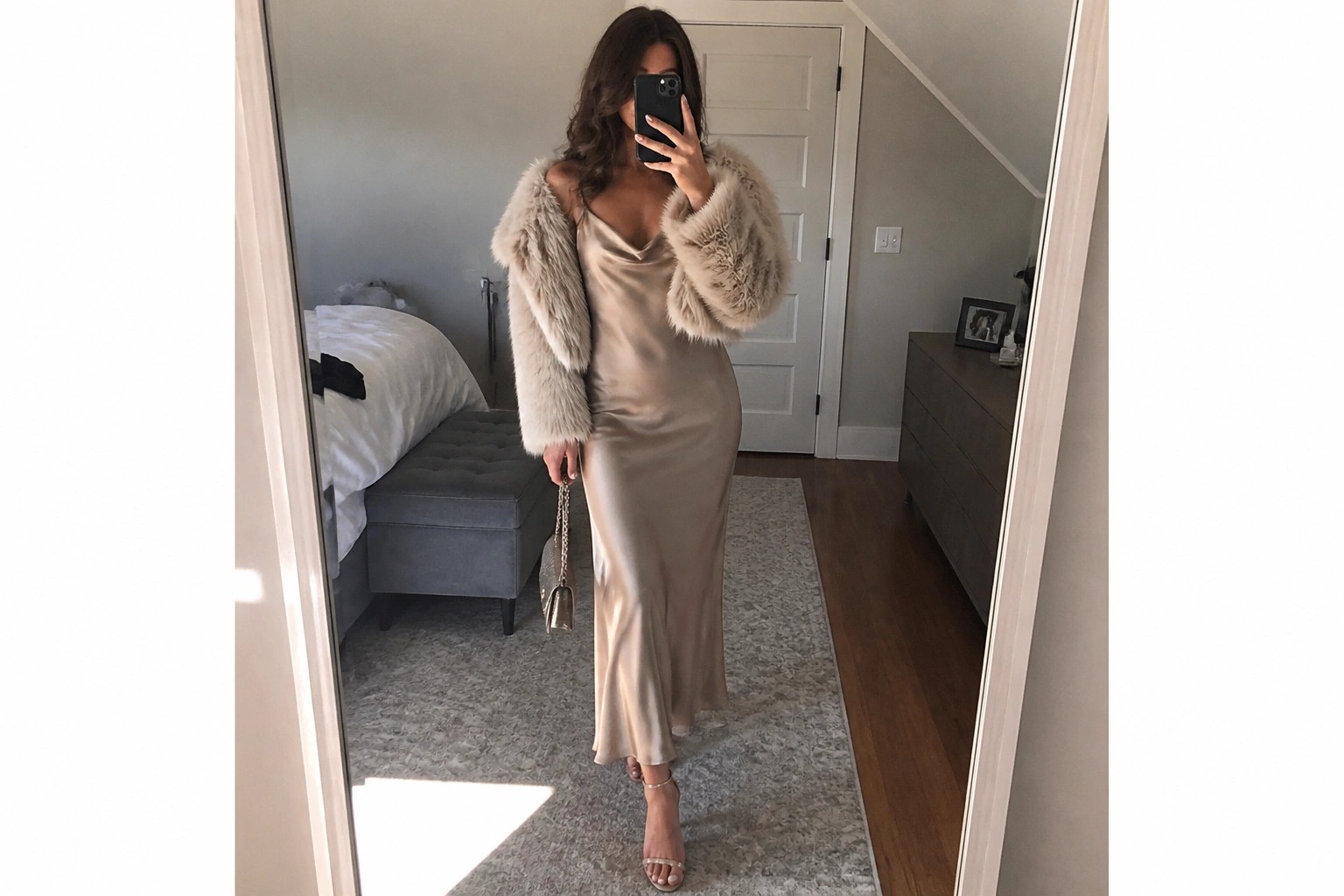 Woman wearing a floor length champagne satin slip dress with V neckline and a short beige fur bolero jacket, holding a small taupe handbag with gold chain strap, standing in front of a mirror.