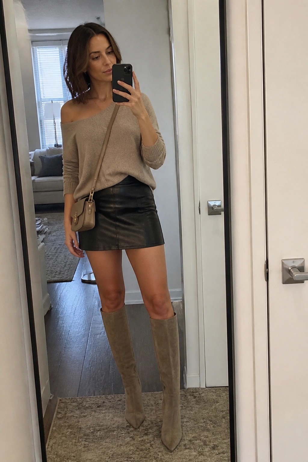 Woman in beige off-shoulder sweater and black leather skirt with suede boots taking mirror selfie