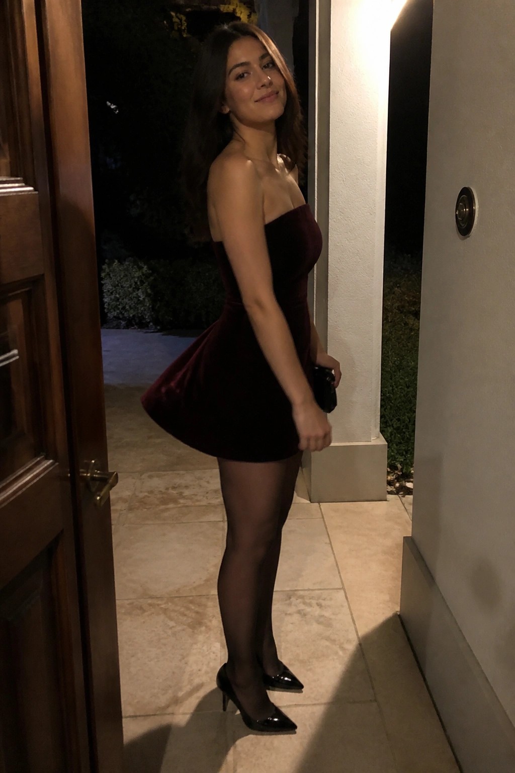 Woman wearing a strapless burgundy velvet mini dress with black sheer tights and black patent pointed toe heels while holding a small black clutch.