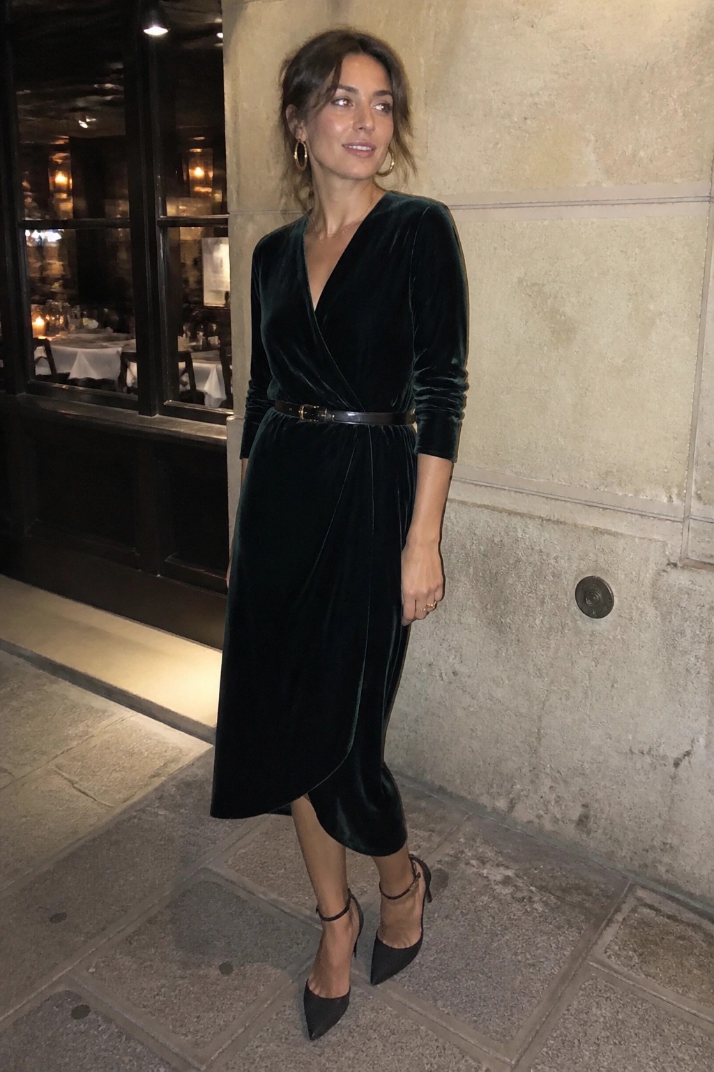 Woman in dark green velvet wrap dress and heels outside restaurant at night
