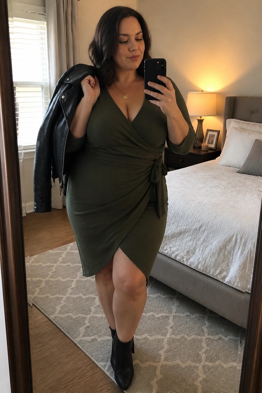 Woman in olive wrap dress taking mirror selfie with leather jacket in bedroom