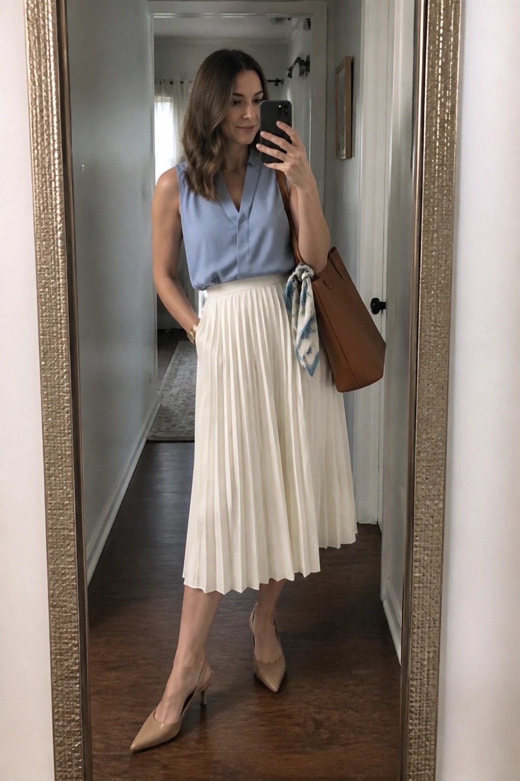Woman in blue sleeveless top and white pleated skirt taking mirror selfie with brown bag