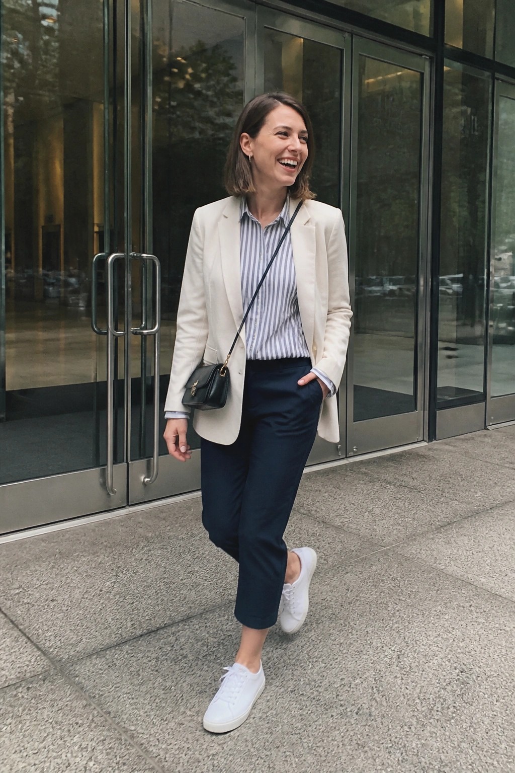 Woman in cream blazer, striped shirt, navy pants, and white sneakers before glass doors