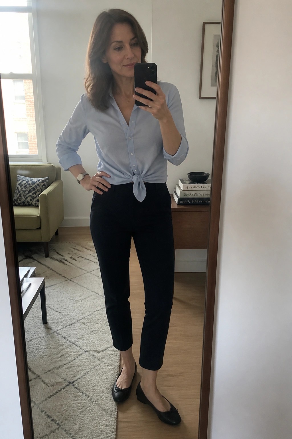Woman in light blue tied blouse and black pants taking mirror selfie
