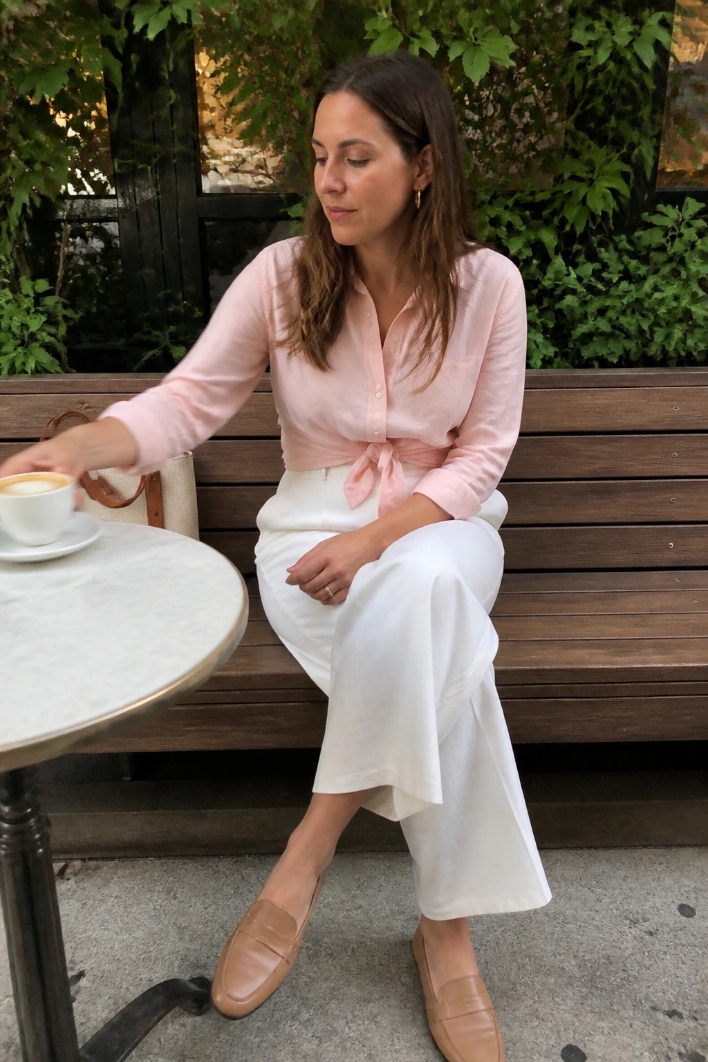 Woman in pink tied shirt and white pants reaching for coffee on outdoor bench.