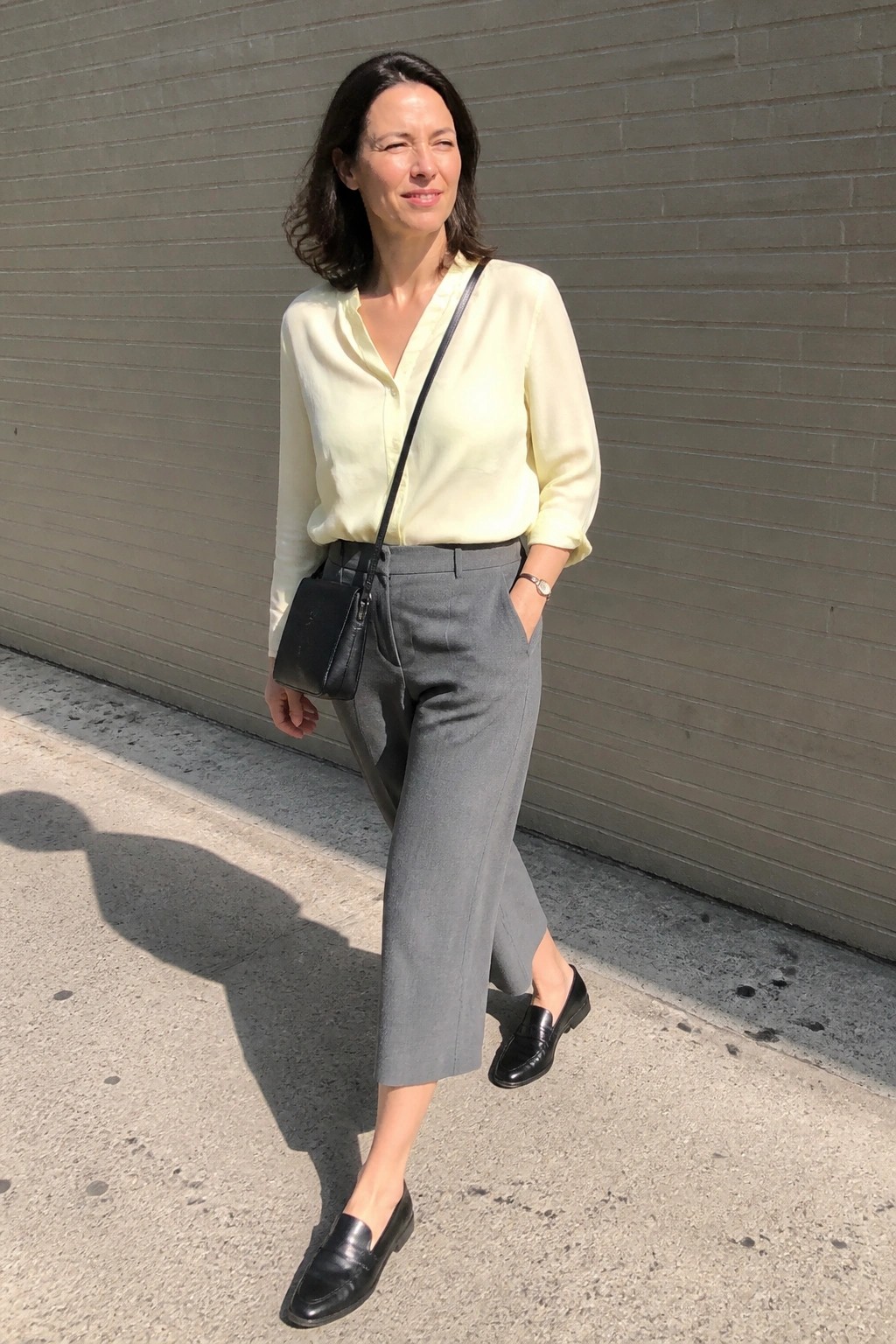 Woman in pale yellow blouse and gray trousers winking by brick wall.