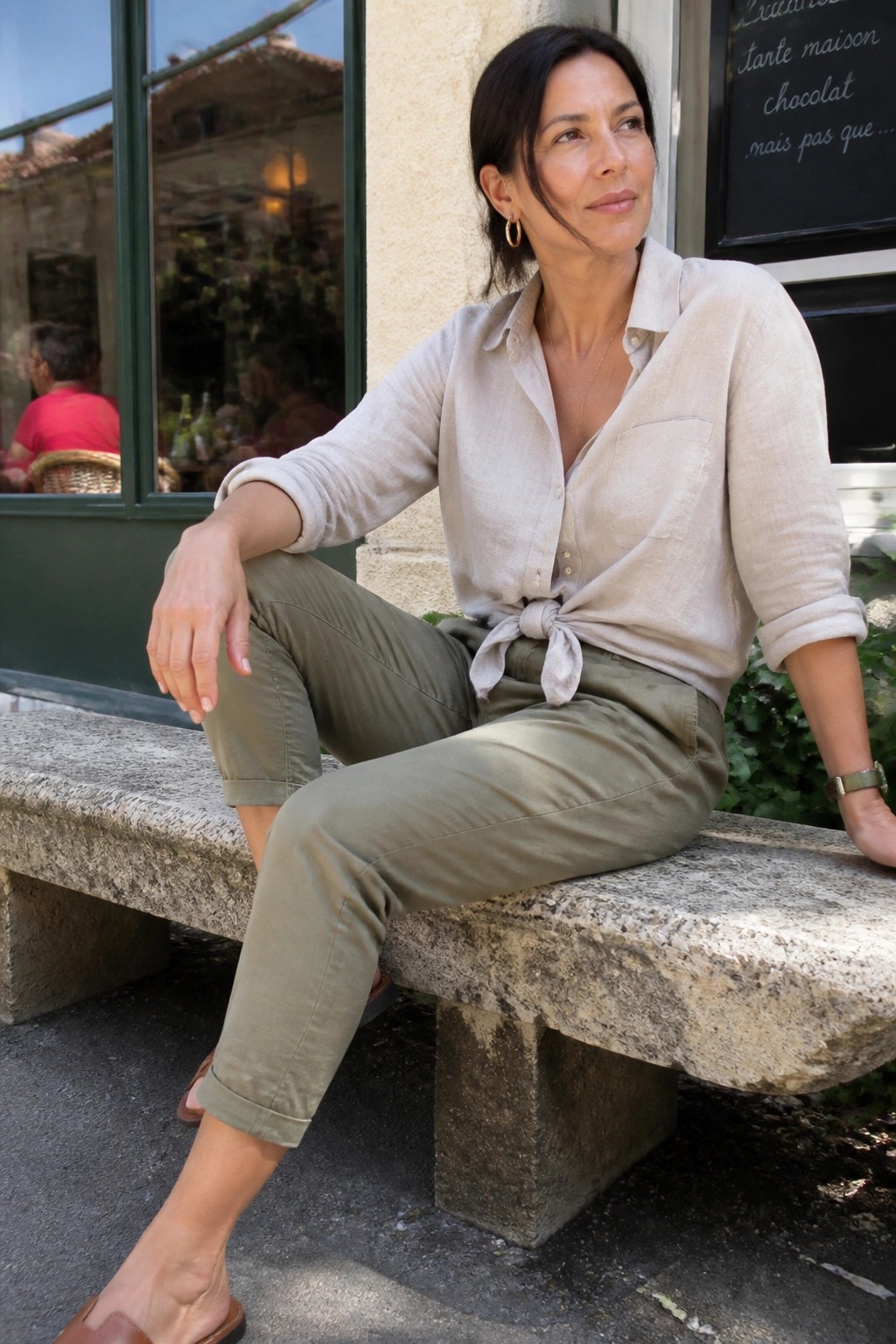 Woman in beige tied shirt and olive pants seated on stone bench outside cafe