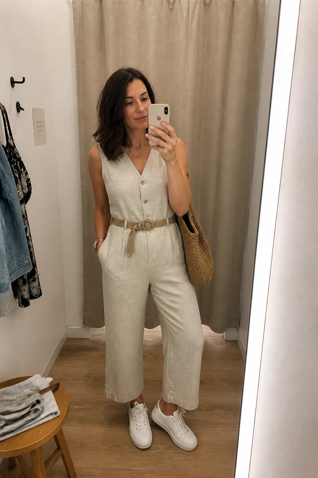 Woman in beige linen jumpsuit takes mirror selfie in fitting room with woven bag