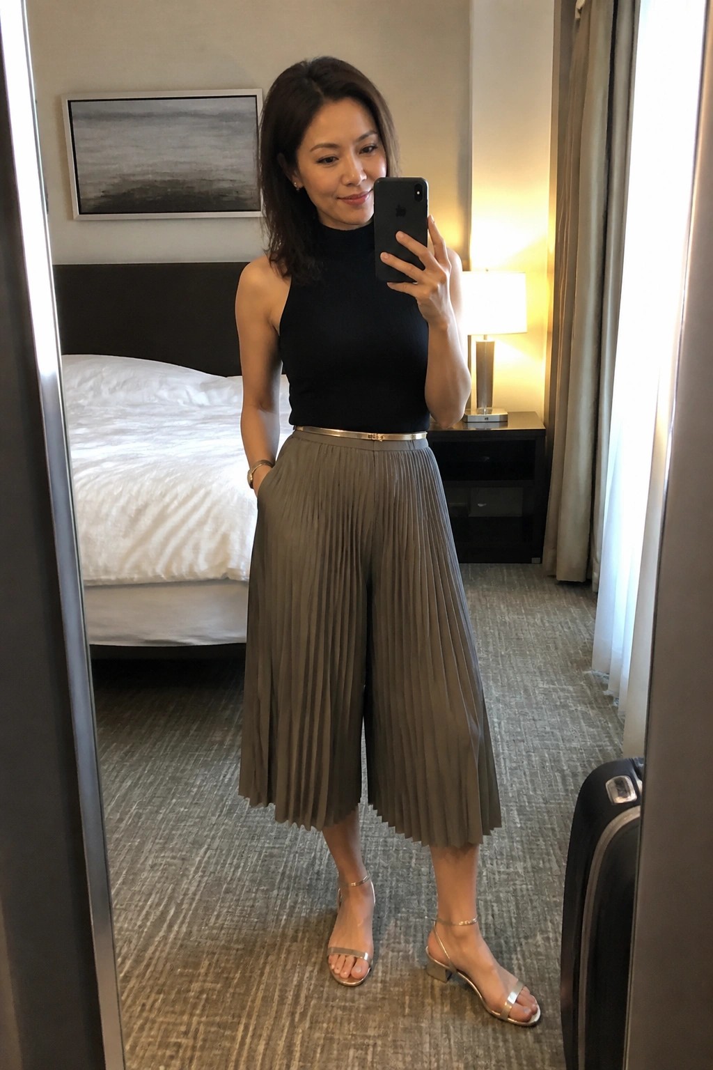 Woman in black top and pleated olive pants taking mirror selfie in hotel room