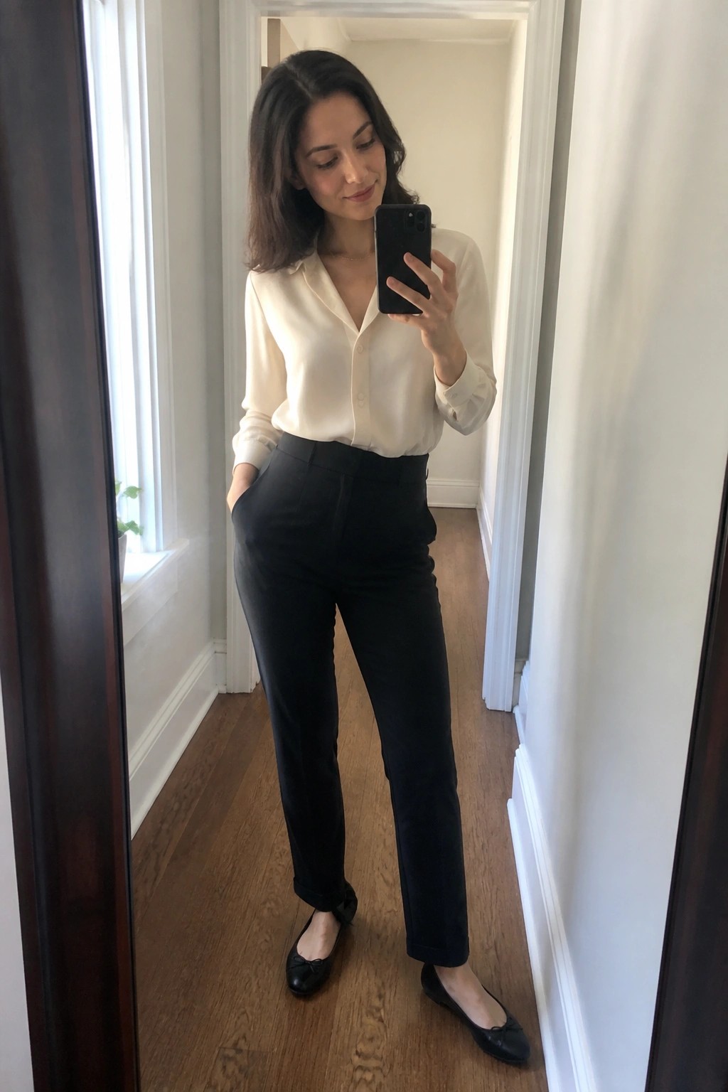 Woman in cream blouse and black pants taking mirror selfie with phone