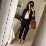 Woman in navy blazer and black pants taking mirror selfie with brown tote bag