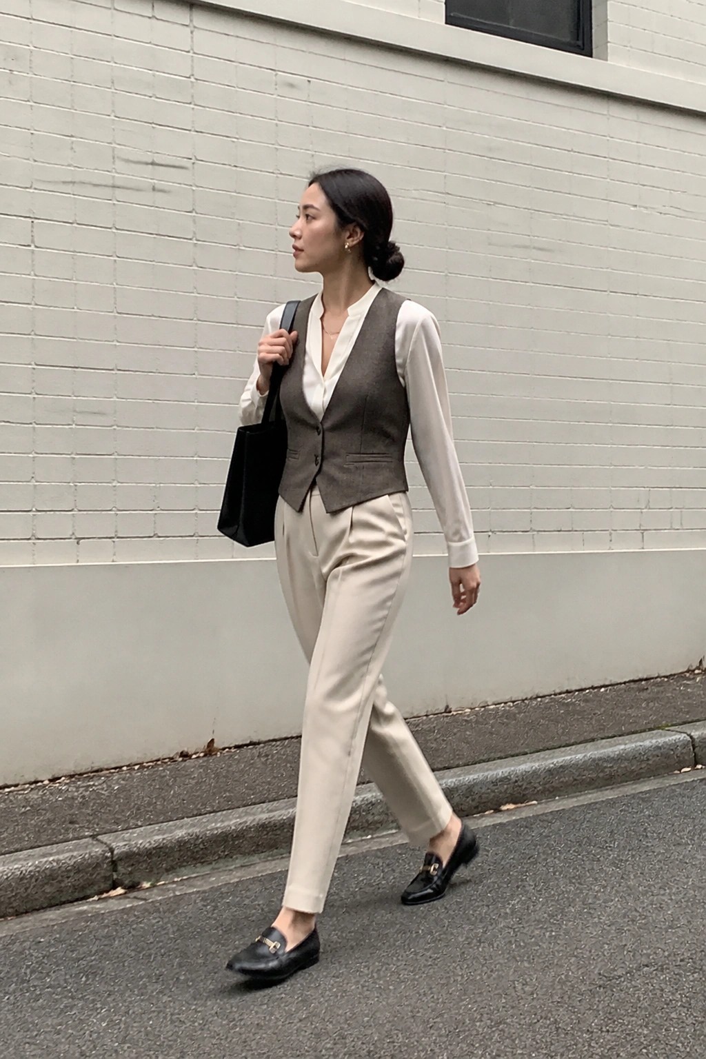 Woman in brown vest and beige trousers carrying black bag on city street