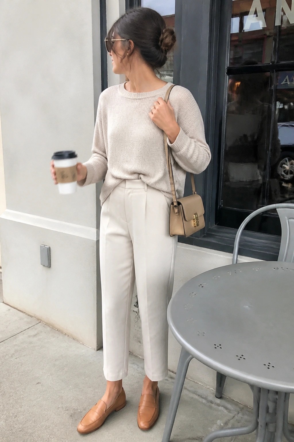 Woman in beige knit sweater and trousers holding coffee with crossbody bag