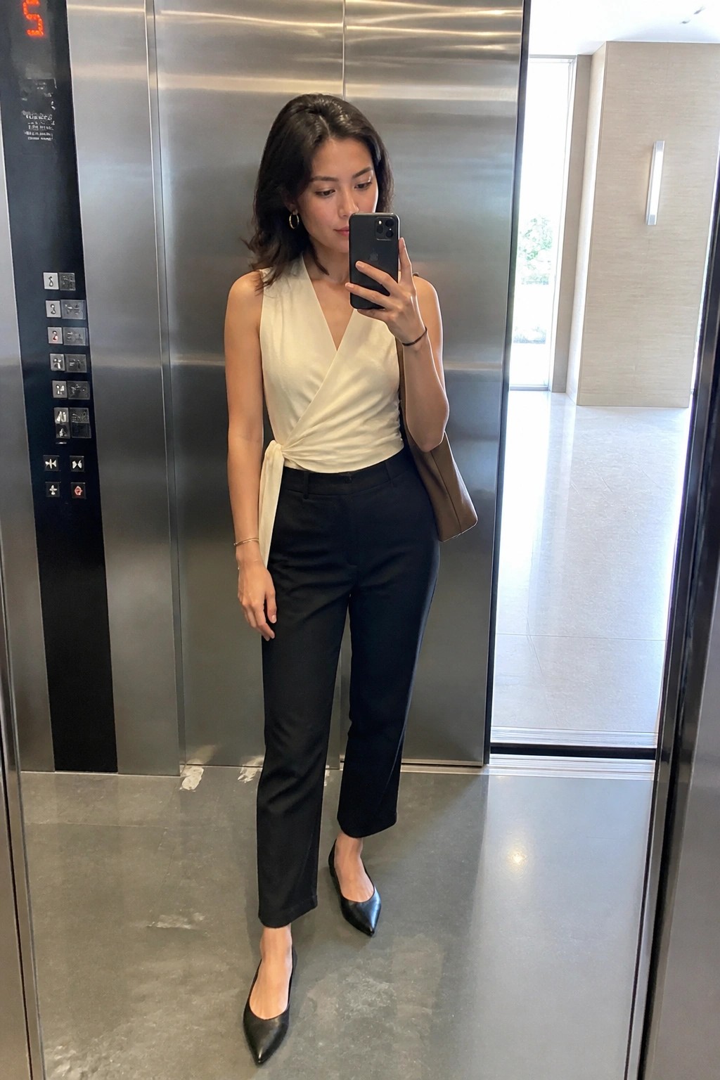 Woman wearing cream sleeveless wrap blouse tucked into black straight trousers with pointed black flats and brown tote bag while taking a mirror selfie.