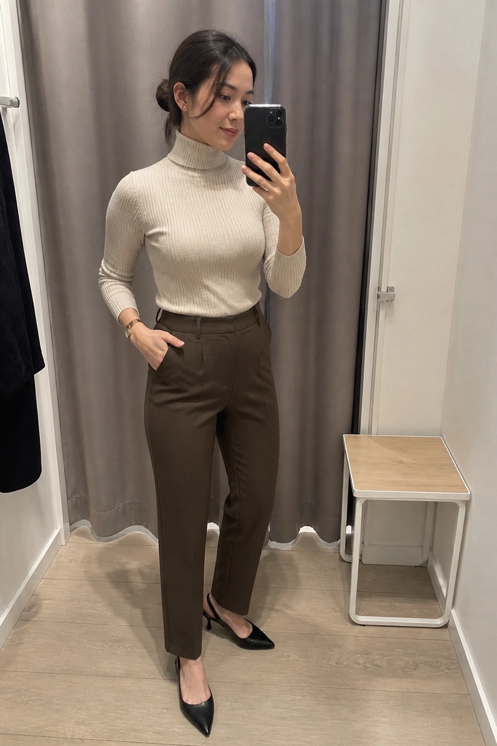 Woman in beige turtleneck and brown pants taking mirror selfie in fitting room