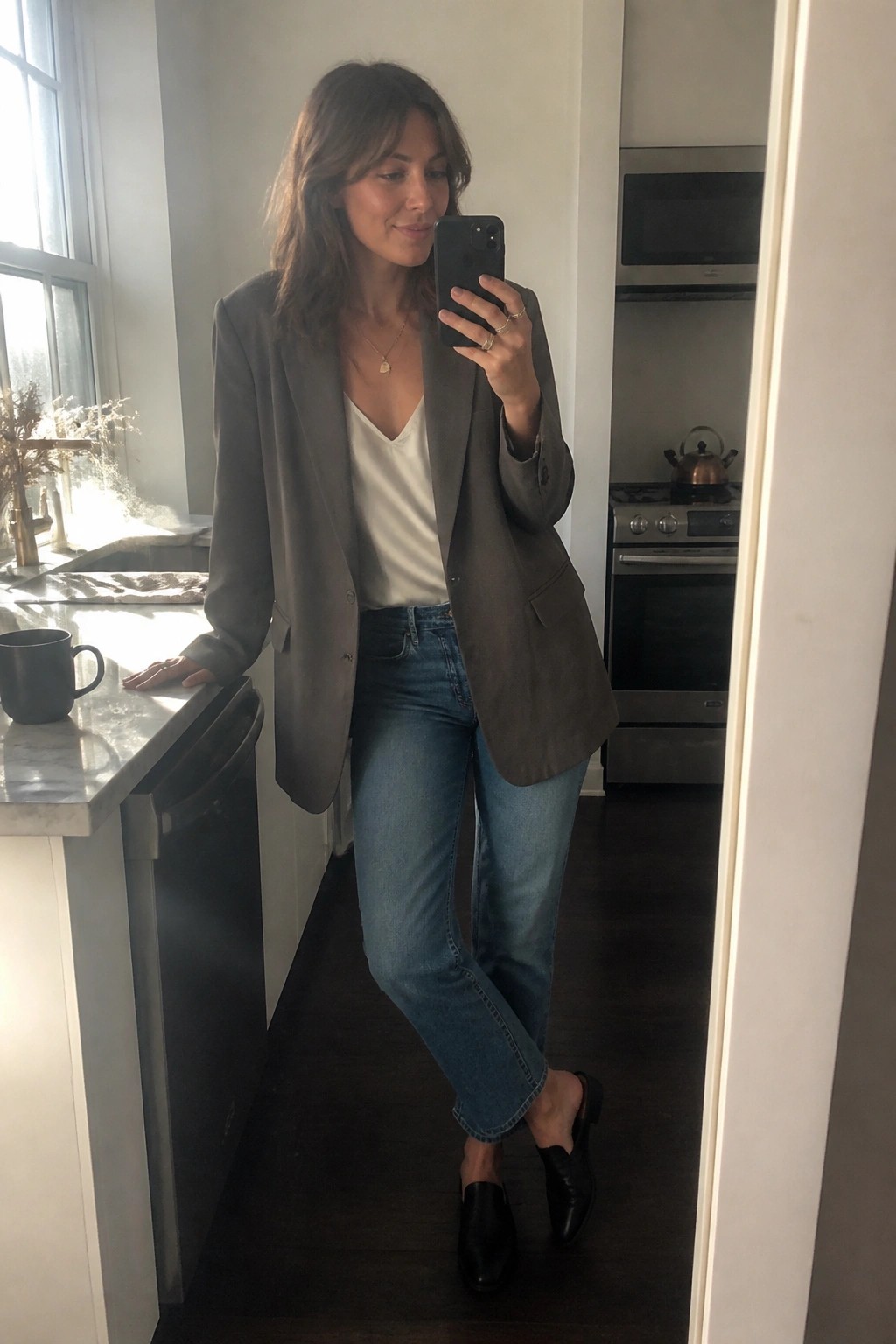Woman in taupe blazer and jeans taking mirror selfie in kitchen
