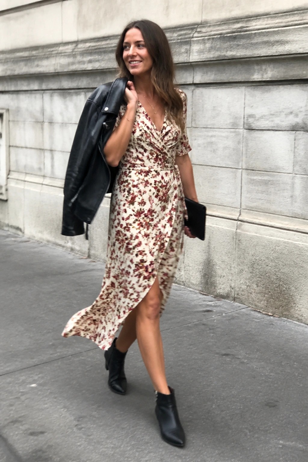 Woman in floral wrap dress carrying black leather jacket on city sidewalk