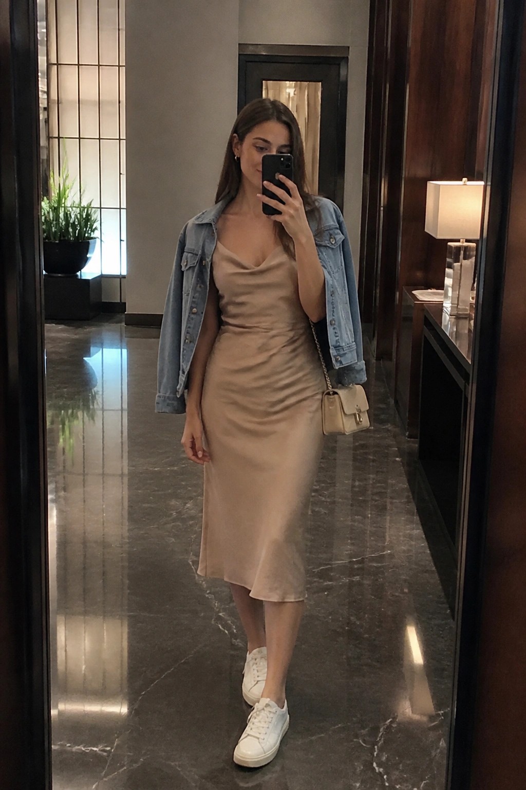 Woman in beige slip dress and denim jacket taking mirror selfie in luxury hallway