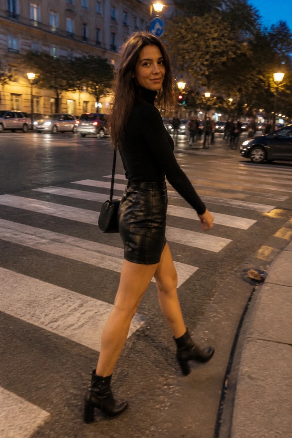 Woman wearing black turtleneck top tucked into black leather mini skirt with black ankle boots and small crossbody bag while walking on a city crosswalk.