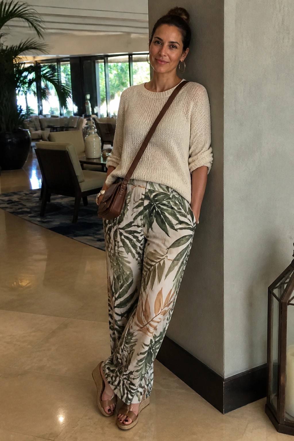 Woman standing indoors wearing a cream knit sweater with rolled sleeves tucked into wide leg pants featuring green and brown tropical leaf prints, brown leather crossbody bag, and brown wedge sandals.