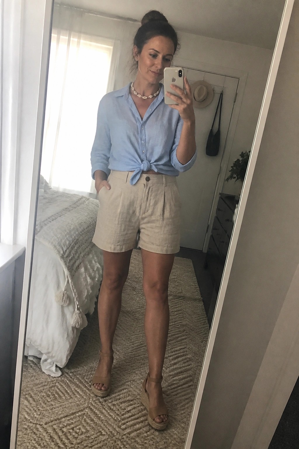 Woman wearing light blue button down shirt tied at waist with beige linen shorts, shell necklace, and tan wedge sandals in mirror selfie pose.