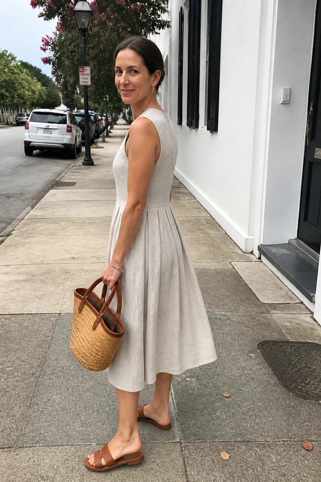 Woman in light beige sleeveless midi linen dress with pleated skirt and open back detail, carrying a straw basket tote with brown leather handles, wearing brown slide sandals and delicate gold jewelry.