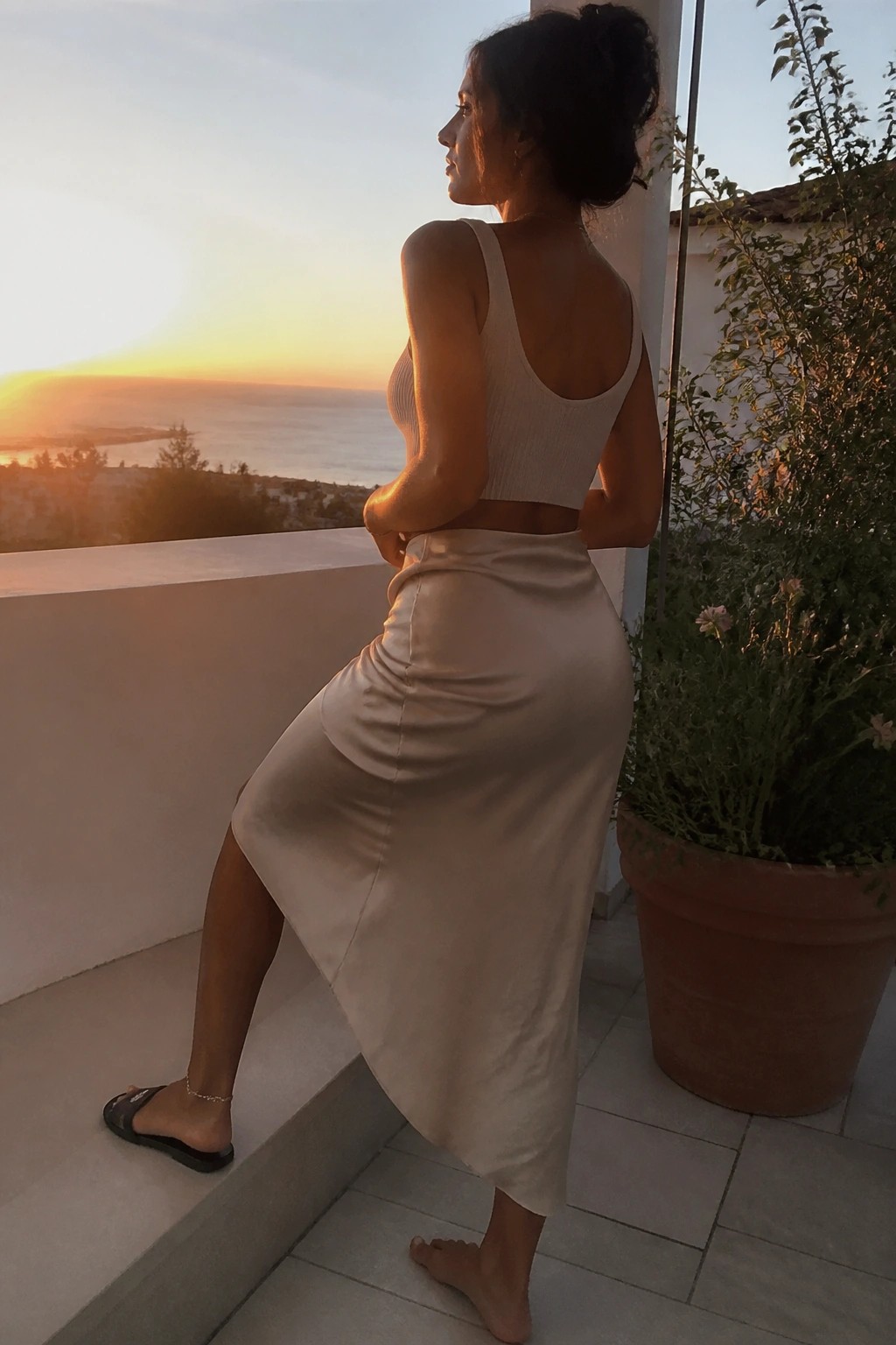Woman in beige crop top and satin skirt on ocean-view balcony at sunset