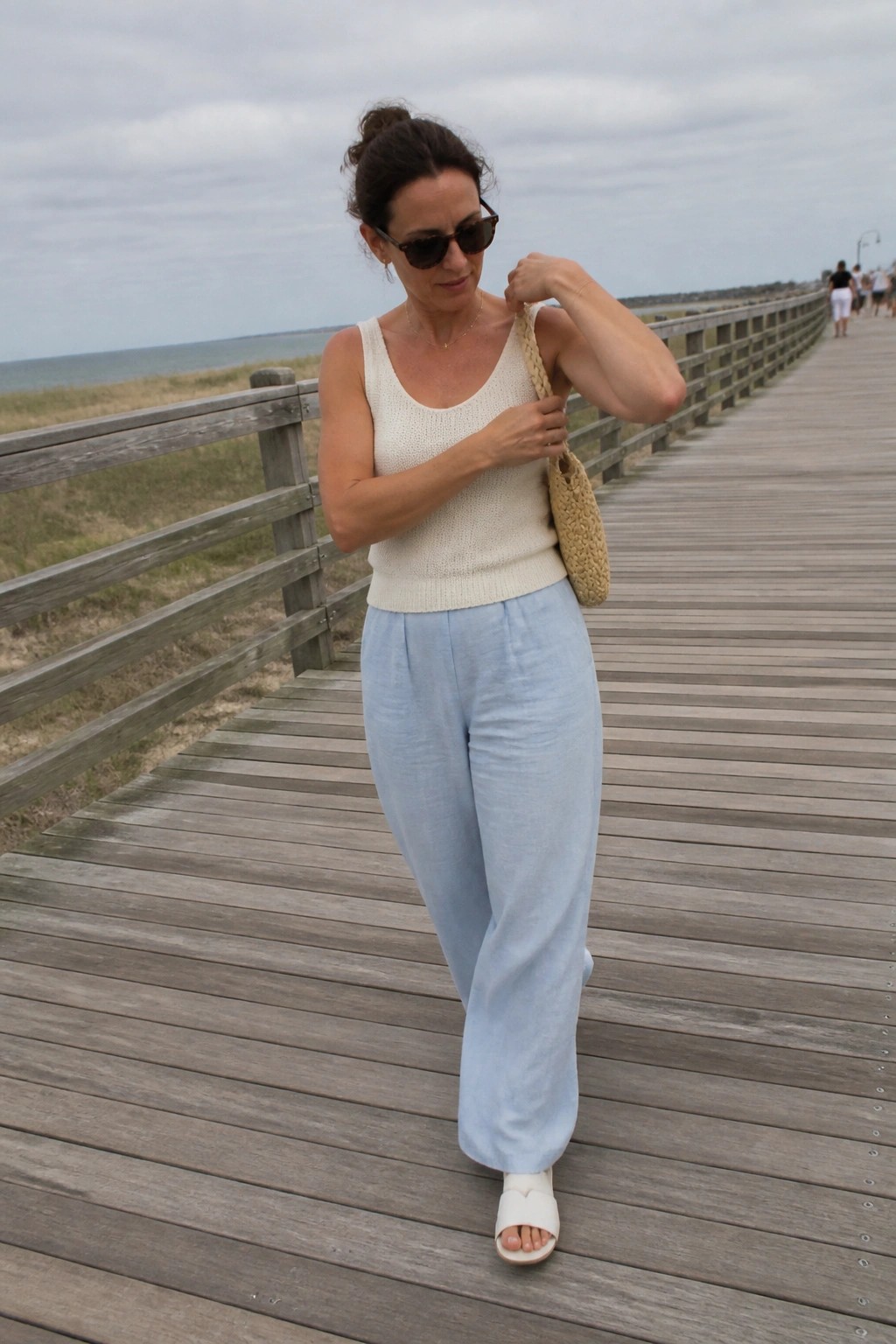 Woman in cream knit tank top and light blue wide-leg linen pants with white slide sandals and woven shoulder bag standing on wooden boardwalk.