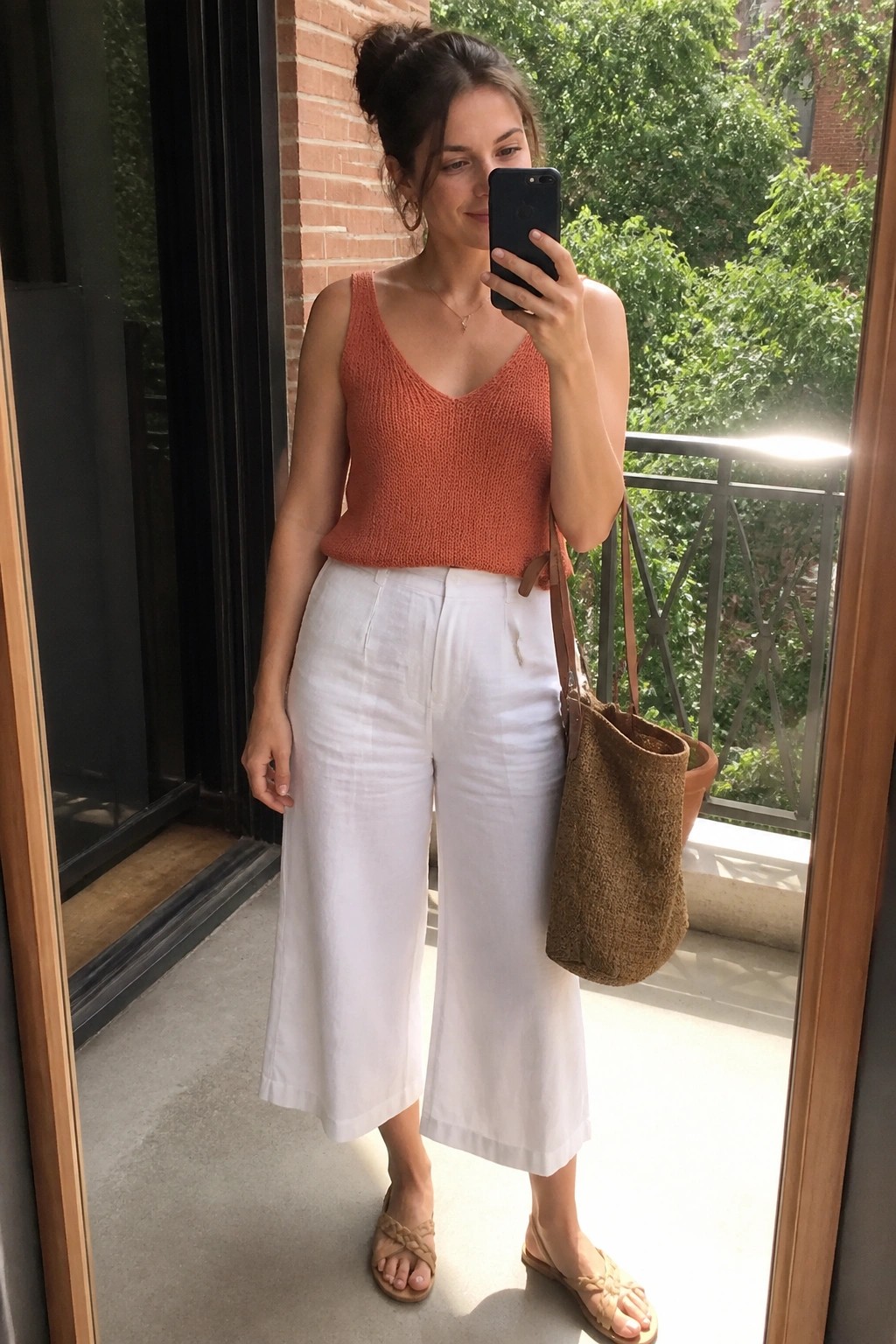 Woman wearing an orange sleeveless knit V-neck tank top tucked into white wide-leg cropped pants, carrying a brown woven shoulder bag and wearing beige braided flat sandals.