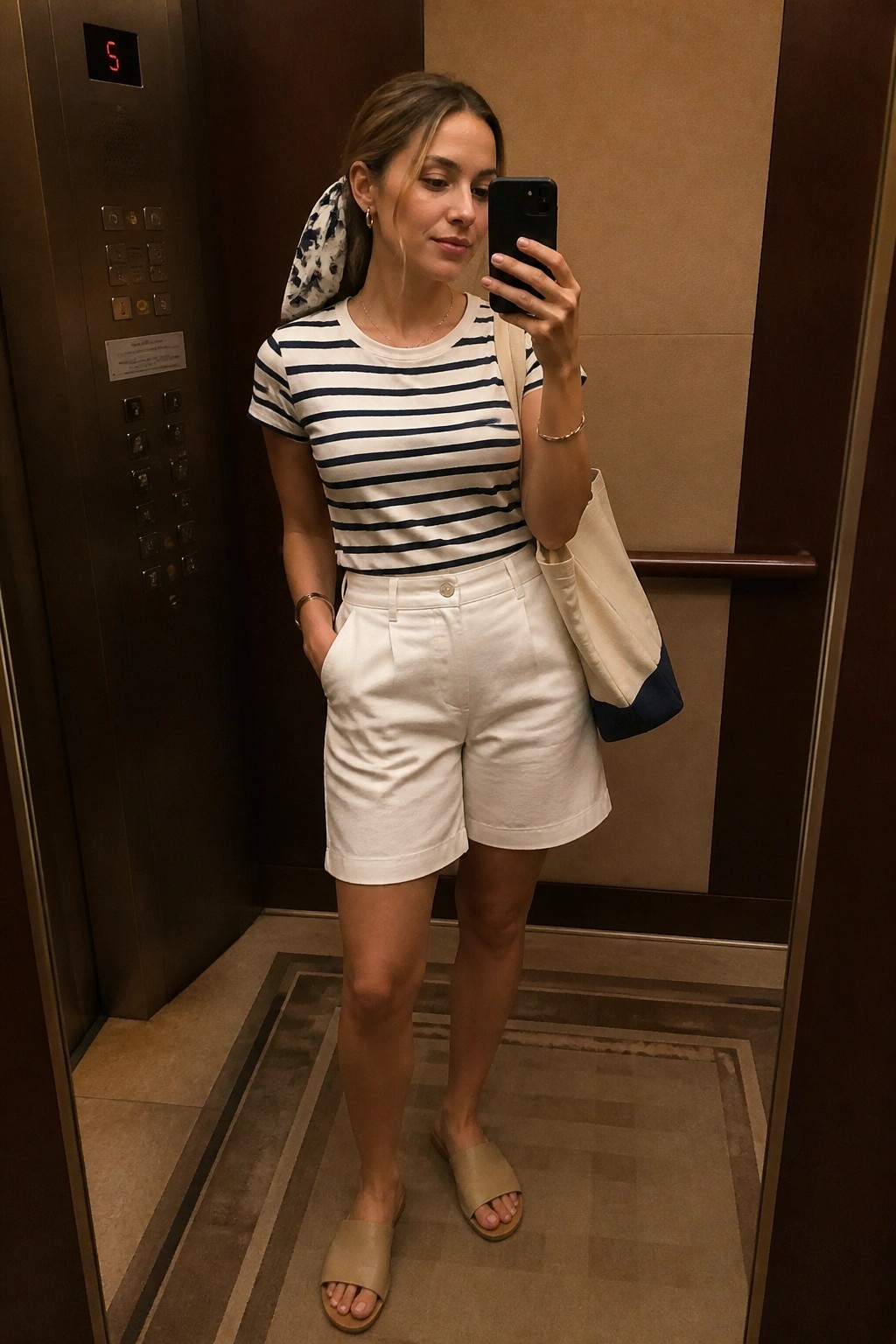 Woman in striped shirt and white shorts taking selfie in elevator with tote bag.
