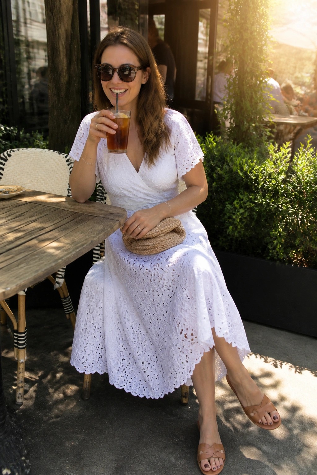 Woman wearing a white eyelet lace wrap midi dress with short flutter sleeves and scalloped hem paired with brown leather slide sandals and holding a woven straw clutch.