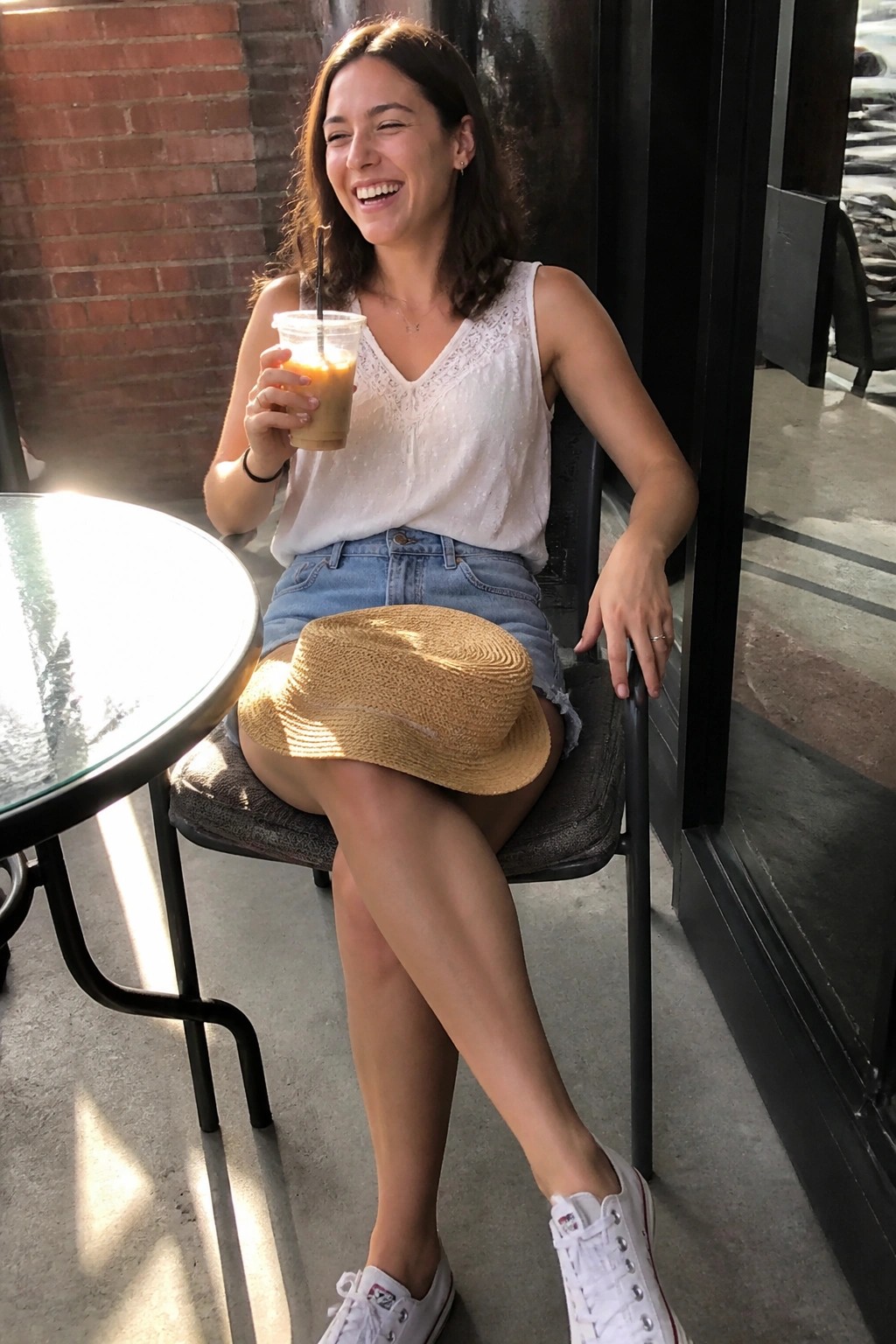 Smiling woman in white top and denim shorts holds iced coffee, straw hat on lap.