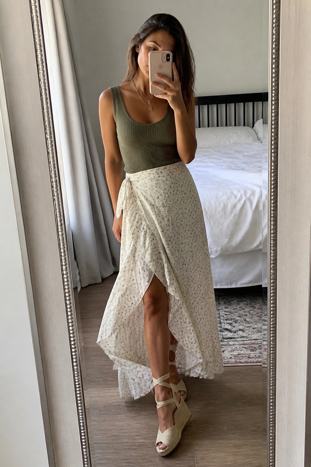 Woman wearing green ribbed tank top with cream floral wrap skirt and beige wedge espadrilles taking a mirror selfie.