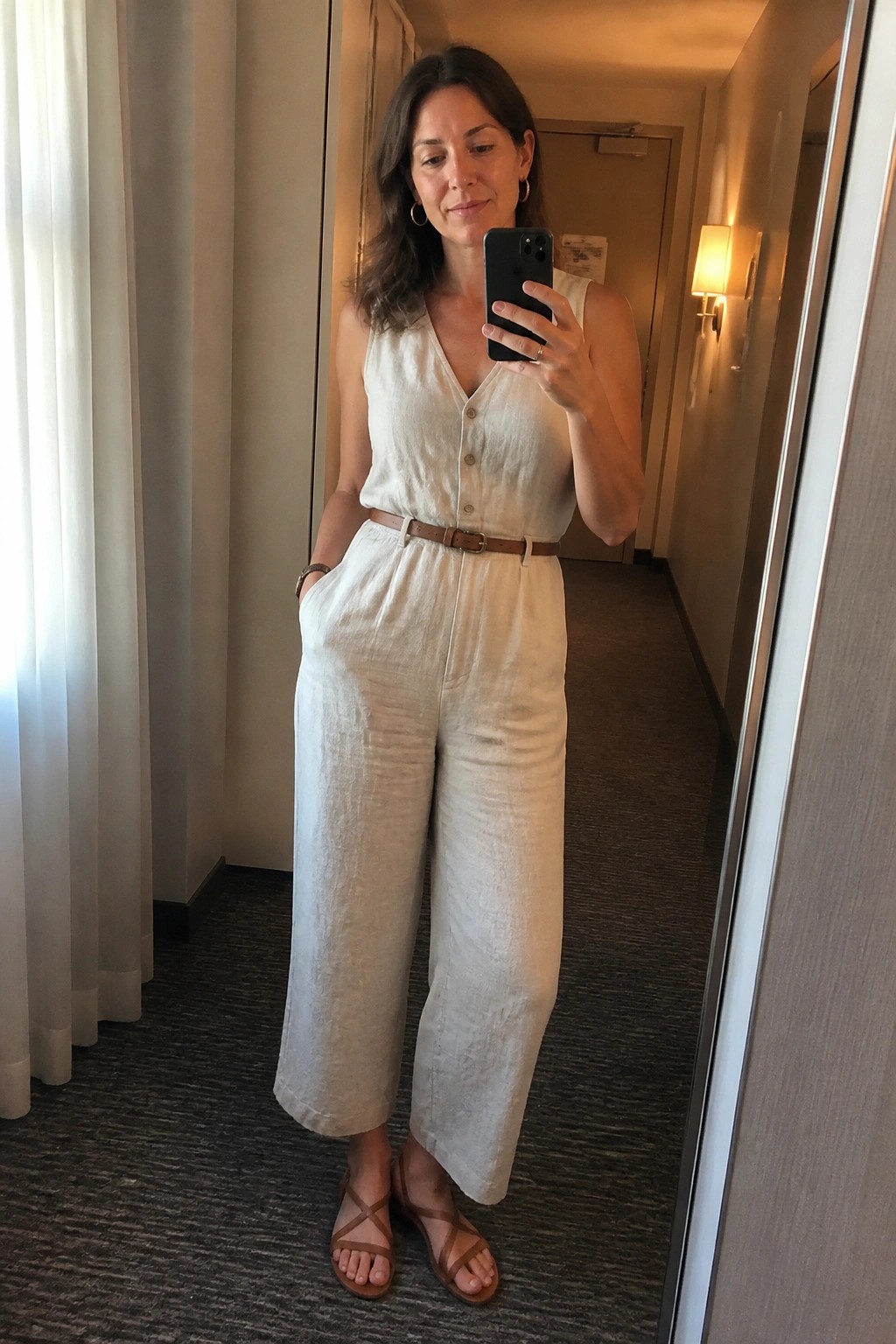 Woman in white linen jumpsuit taking mirror selfie in hallway
