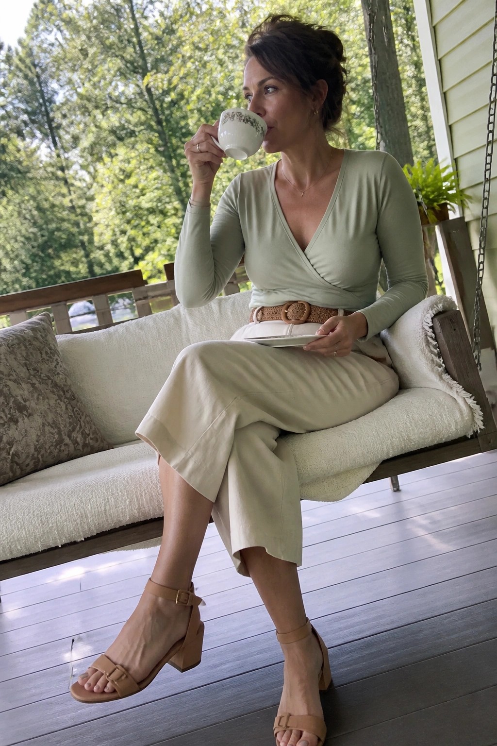 Woman wearing a sage green long sleeve wrap top tucked into cream wide leg culottes with a brown woven belt and beige block heel sandals while holding a teacup on a porch swing.