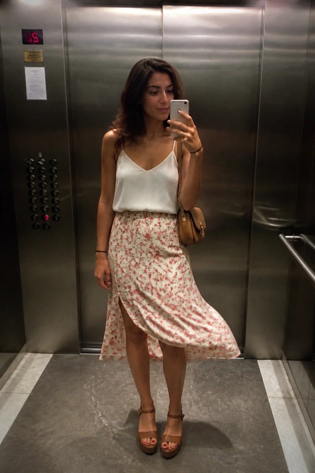 Woman in white camisole and red floral skirt takes selfie in elevator.