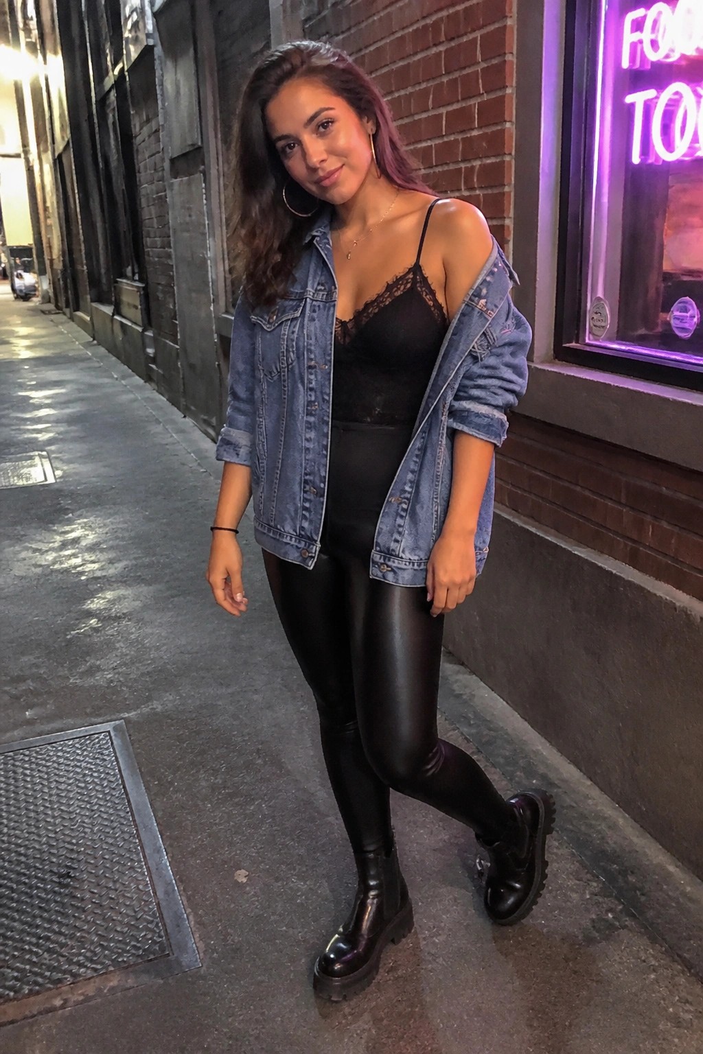 Woman in black lace top, shiny leggings, and denim jacket on nighttime sidewalk