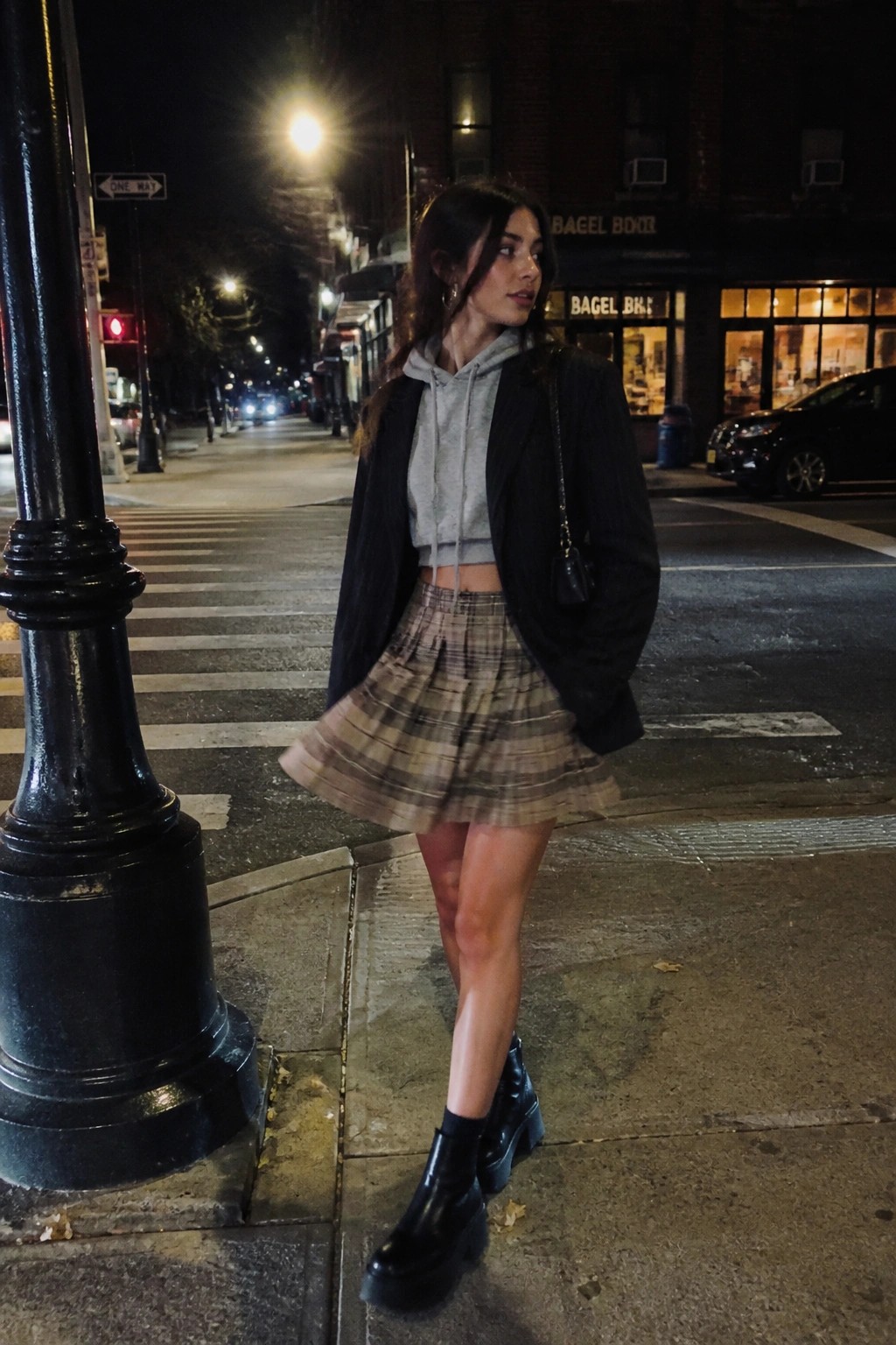Woman in grey hoodie, black blazer, plaid skirt, and boots on city sidewalk at night