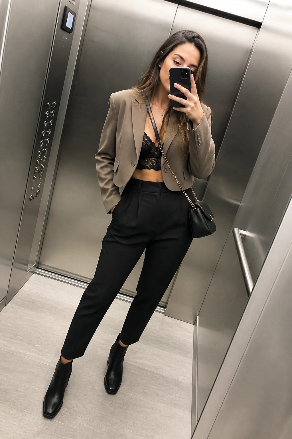 Woman in taupe blazer and black lace top taking elevator mirror selfie